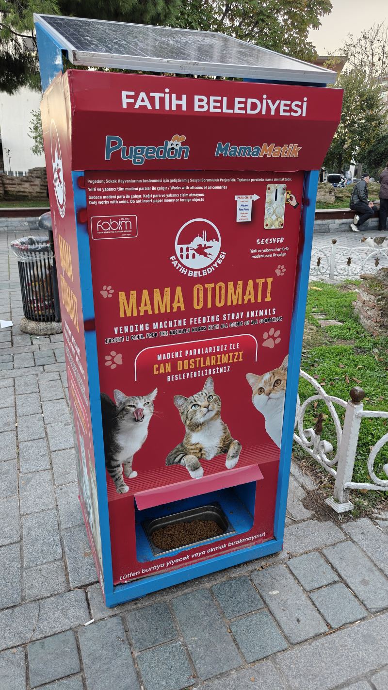 Ein roter Futterautomat der Bezirksverwaltung Fatih für Straßentiere auf einem Gehweg, davor eine Wasserschale