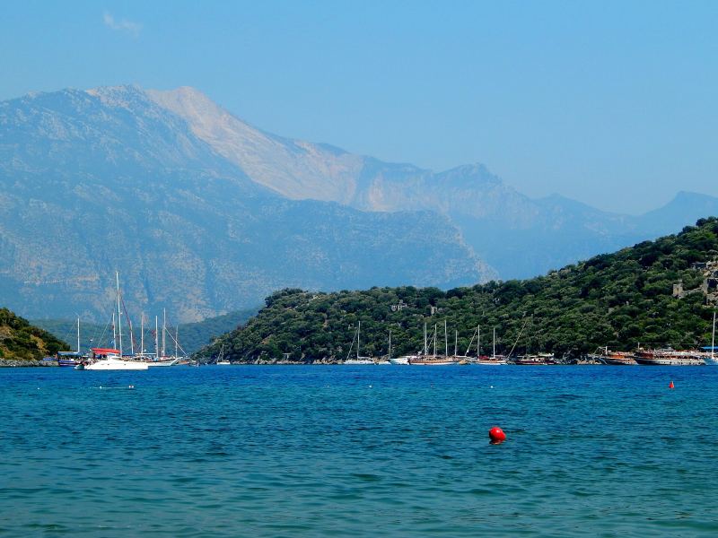 Yachts at anchor off a wooded shore at Gemiler, with high mountains behind